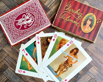 May include: A deck of playing cards with a red box featuring white filigree and a central illustration, alongside a box labelled Cherchez la femme with a portrait. Four cards are fanned out, each displaying a diamond suit and a vintage pin-up illustration.