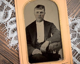 May include: A vintage tintype photograph featuring a portrait of a person with short hair, wearing a dark jacket over a light-coloured shirt. The portrait is encased in a rectangular peach-coloured frame with an arched top and handwritten text at the bottom.
