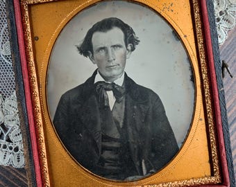May include: A vintage daguerreotype portrait of a person with dark hair, wearing a dark coat, waistcoat, and a bow tie. The photograph is encased in a rectangular black frame with a gold-toned oval mat and a red velvet interior lining.