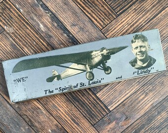 May include: A rectangular metal tin featuring a vintage graphic of the Spirit of St. Louis aeroplane. The text reads "WE", "The Spirit of St. Louis", and "Lindy" alongside a portrait of a smiling aviator. The background is a muted grey tone.