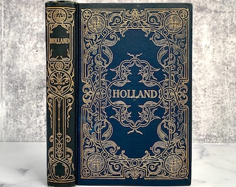 May include: A dark blue hardback book titled Holland with intricate gold foil filigree patterns on the front cover and spine. The spine also features the text Amicis. The design includes symmetrical scrollwork and decorative borders.