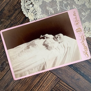 May include: Vintage sepia-toned photograph of a sleeping baby, dressed in a white gown, resting on a white cloth. The photograph has a pink border and the text "Bergstroms Waco, Tex." is printed on the right side.