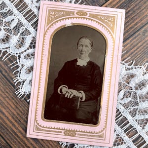 May include: A vintage rectangular photograph in a pink and gold decorative frame featuring a black and white portrait of a person wearing a dark long-sleeved garment with a white collar and cuffs, holding a small rectangular book in their lap.