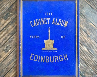 May include: A vintage hardback book titled The Cabinet Album Views of Edinburgh with a vibrant blue cover and gold embossed lettering. The front cover features a central gold illustration of the Old Mercat Cross monument in Edinburgh.