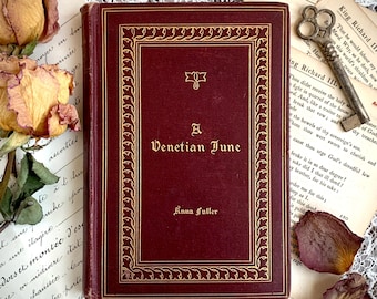 May include: A maroon hardback book titled A Venetian June by Anna Fuller with gold embossed lettering and a decorative border. The book is surrounded by dried roses, a vintage metal skeleton key, and pages of antique printed text.