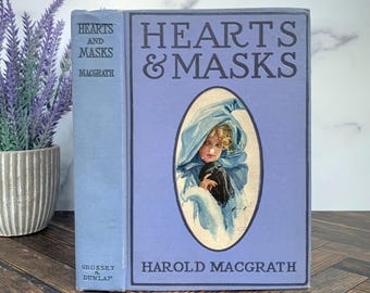 May include: A light blue hardback book titled Hearts & Masks by Harold MacGrath. The front cover features a central oval illustration of a person wearing a blue hooded cloak and holding a black mask. The spine displays the title and author.