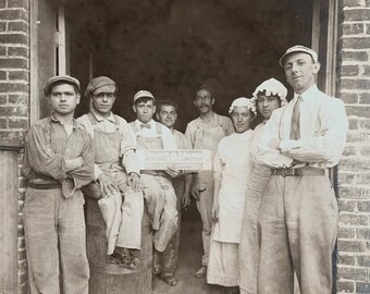 May include: A black and white photograph of eight individuals standing in a brick doorway. One person holds a sign that reads "Cosmopolitan Magazine". The group wears early 20th-century work attire, including caps, aprons, overalls, and trousers.