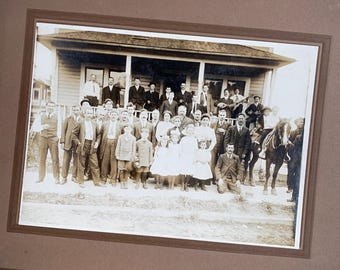 May include: A vintage sepia-toned photograph in a brown mat frame depicting a large group of people posed in front of a wooden house with a porch. The group includes adults in suits and hats, children in light-coloured clothing, and one horse.