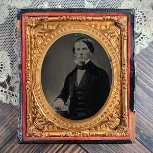 May include: Antique framed portrait of a man in formal attire. The oval portrait is set within an ornate gold-colored frame, with a red border. The man is wearing a dark suit and vest, with a white collared shirt and a dark bow tie.