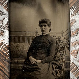 May include: A vintage sepia-toned photograph featuring a person with dark hair styled in a bun, wearing a high-collared, long-sleeved dark garment with intricate embroidery. The person is seated on a patterned sofa with a floral design.