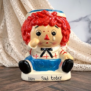 May include: A ceramic doll-shaped container with red hair, a blue apron, and a black bow tie. The doll has a sad expression with a tear and the text "I am Sad today" on the base. The doll is sitting down.