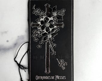May include: A rectangular black booklet with a silver embossed cross and floral design on the cover. The text "OFFRANDES DE MESSES" is printed in silver at the bottom. A thin black cord with a tassel is attached to the left side of the cover.