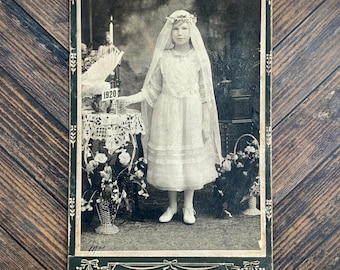 May include: A black and white vintage photograph of a child in a white dress and veil, standing beside a table with a candle and a sign reading 1920. The bottom border features the text Urbanowicz 4852 Ashland Ave Chicago.