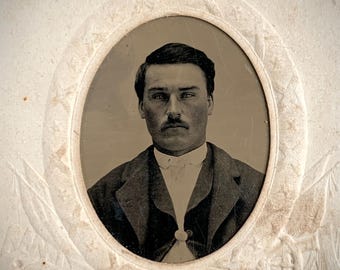 May include: A vintage sepia-toned portrait photograph of a person with short dark hair and a moustache, wearing a collared shirt and a dark jacket. The photograph is encased in an oval frame featuring embossed decorative details of flags and wheels.