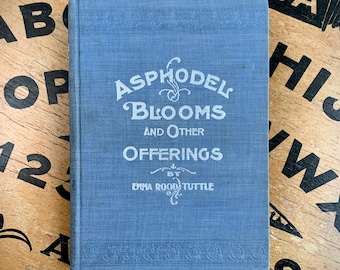 May include: A blue hardback book titled Asphodel Blooms and Other Offerings by Emma Rood Tuttle. The front cover features white embossed lettering and decorative scrollwork, with a textured fabric finish and a horizontal band pattern at the base.