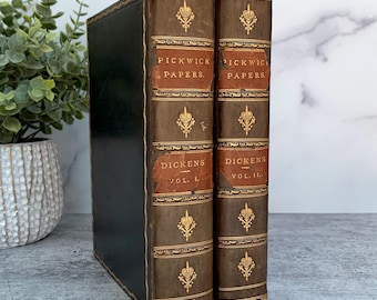 May include: Two antique books titled Pickwick Papers by Dickens, Volume I and Volume II. The books feature dark brown leather spines with gold-embossed crown motifs and lettering, set against a black cover. The spines show signs of wear.