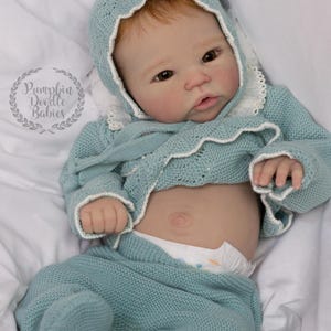 May include: A baby doll wearing a light blue knitted outfit, including a bonnet, sweater, and pants. The outfit has white trim. The doll has fair skin, red hair, and is lying on a white surface. The doll is wearing a diaper.
