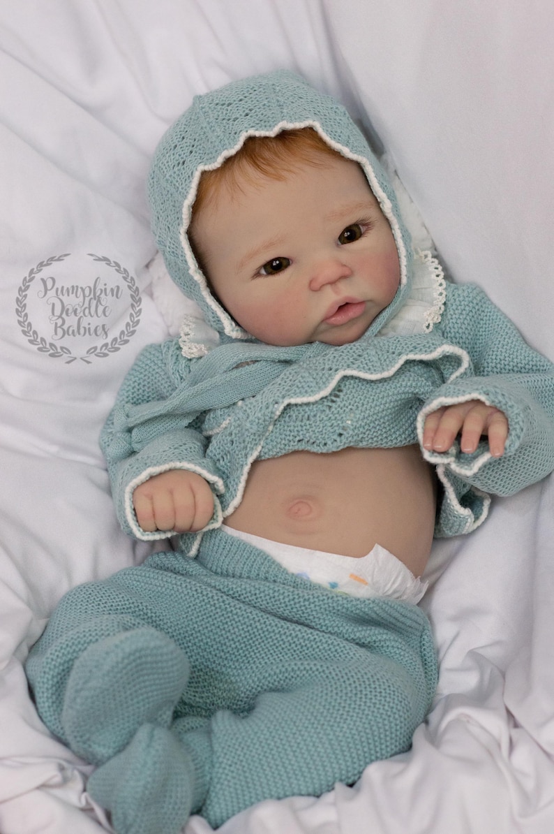 May include: A baby doll wearing a light blue knitted outfit, including a bonnet, sweater, and pants. The outfit has white trim. The doll has fair skin, red hair, and is lying on a white surface. The doll is wearing a diaper.
