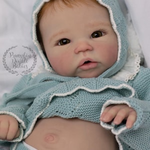 May include: A realistic baby doll with fair skin, red hair, and brown eyes. The doll wears a light blue knitted outfit with a matching bonnet trimmed with white lace. The doll's stomach is visible, and it wears a diaper. The logo "Pumpkin Doodle Babies" is in the upper left.