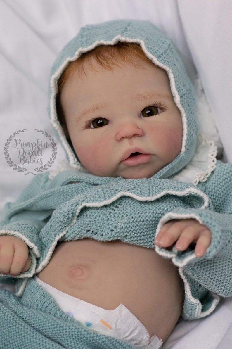 May include: A realistic baby doll with fair skin, red hair, and brown eyes. The doll wears a light blue knitted outfit with a matching bonnet trimmed with white lace. The doll's stomach is visible, and it wears a diaper. The logo "Pumpkin Doodle Babies" is in the upper left.
