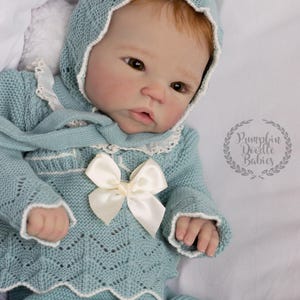 May include: A baby doll wearing a light blue knitted outfit, including a bonnet, sweater, and pants. The sweater features a white bow and trim. The doll has red hair and is lying on a white surface. The image includes the text "Pumpkin Doodle Babies".