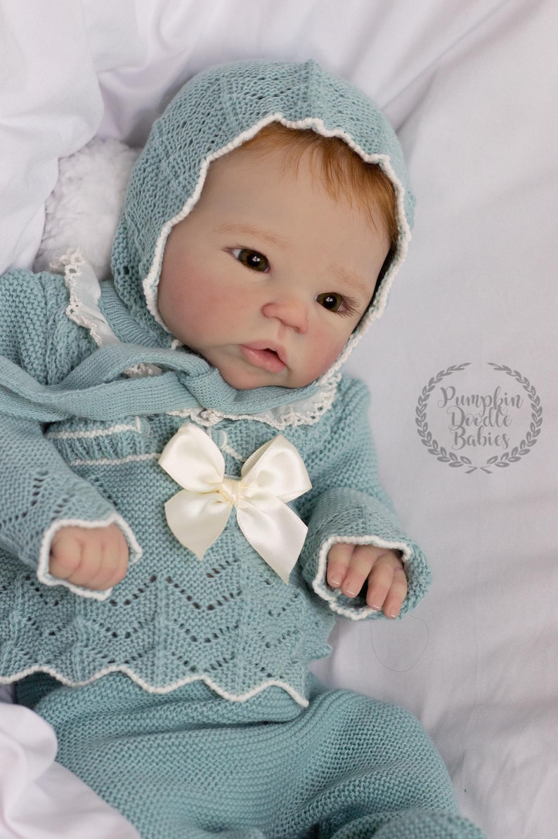 May include: A baby doll wearing a light blue knitted outfit, including a bonnet, sweater, and pants. The sweater features a white bow and trim. The doll has red hair and is lying on a white surface. The image includes the text "Pumpkin Doodle Babies".