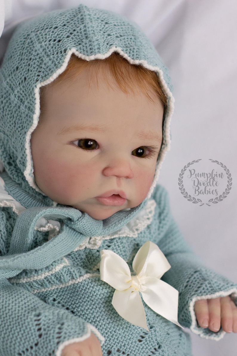May include: A baby doll wearing a light blue knitted outfit with a matching bonnet. The bonnet and outfit are trimmed with white lace. A large white bow adorns the front of the outfit. The doll has brown eyes and reddish-blonde hair. The text "Pumpkin Doodle Babies" is visible.