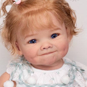 Pode incluir: Um close-up de uma boneca beb&ecirc; com cabelo loiro encaracolado, olhos azuis e bochechas rosadas. A boneca est&aacute; vestindo um vestido branco com uma borda verde e branca xadrez e pompons brancos.