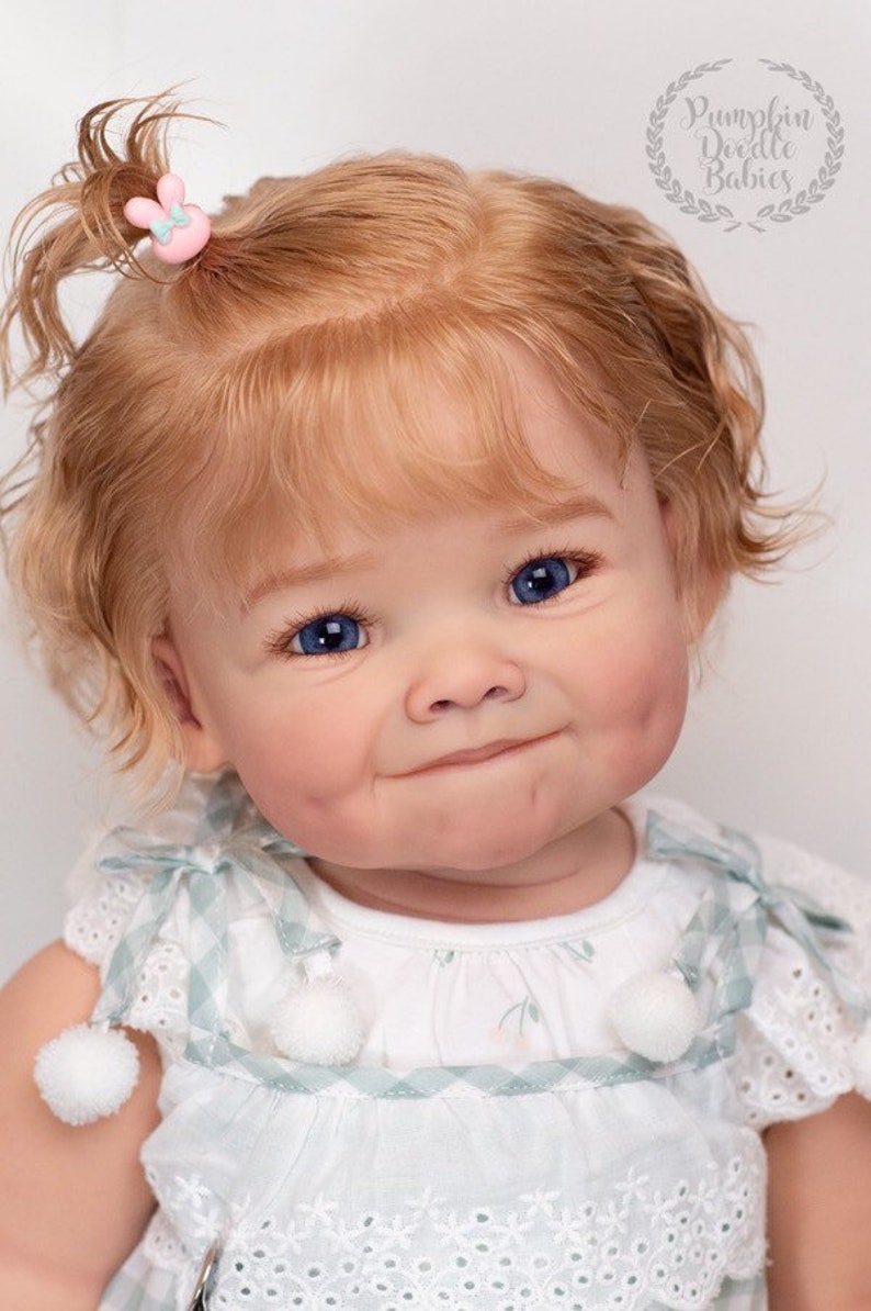 Pode incluir: Um close-up de uma boneca beb&ecirc; com cabelo loiro encaracolado, olhos azuis e bochechas rosadas. A boneca est&aacute; vestindo um vestido branco com uma borda verde e branca xadrez e pompons brancos.