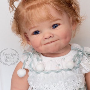 Pode incluir: Um close-up de uma boneca beb&ecirc; com cabelo loiro encaracolado, olhos azuis e bochechas rosadas. A boneca est&aacute; vestindo um vestido xadrez branco e verde com uma borda de renda branca e pompons brancos. A boneca tem um prendedor de cabelo de coelho rosa no cabelo.