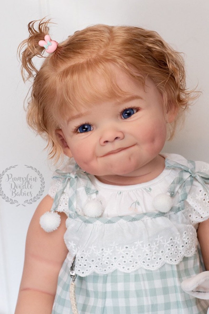 Pode incluir: Um close-up de uma boneca beb&ecirc; com cabelo loiro encaracolado, olhos azuis e bochechas rosadas. A boneca est&aacute; vestindo um vestido xadrez branco e verde com uma borda de renda branca e pompons brancos. A boneca tem um prendedor de cabelo de coelho rosa no cabelo.