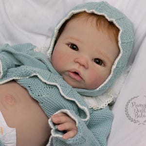 May include: A realistic baby doll wearing a light blue knitted outfit with a matching bonnet. The doll has fair skin, red hair, and brown eyes. The outfit has white trim. The doll is lying on a white surface.