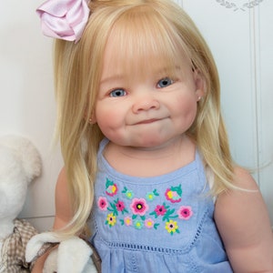 Pode incluir: Um close-up de uma boneca beb&ecirc; com cabelo loiro usando um vestido azul claro com flores bordadas. A boneca tem olhos azuis e um la&ccedil;o rosa no cabelo. A boneca est&aacute; segurando um coelho de pel&uacute;cia branco.