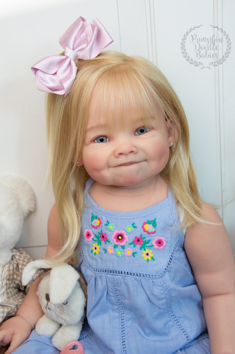 Pode incluir: Um close-up de uma boneca beb&ecirc; com cabelo loiro usando um vestido azul claro com flores bordadas. A boneca tem olhos azuis e um la&ccedil;o rosa no cabelo. A boneca est&aacute; segurando um coelho de pel&uacute;cia branco.
