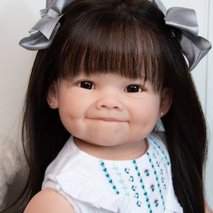 Pode incluir: Um close-up de uma boneca beb&ecirc; com cabelo preto longo, olhos castanhos e um sorriso doce. A boneca est&aacute; vestindo um vestido branco com flores bordadas e tem la&ccedil;os prateados no cabelo.