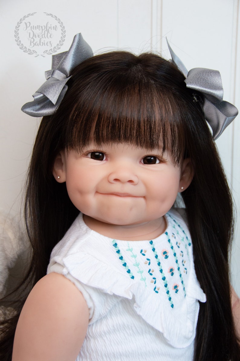 Pode incluir: Um close-up de uma boneca beb&ecirc; com cabelo preto longo, olhos castanhos e um sorriso doce. A boneca est&aacute; vestindo um vestido branco com flores bordadas e tem la&ccedil;os prateados no cabelo.