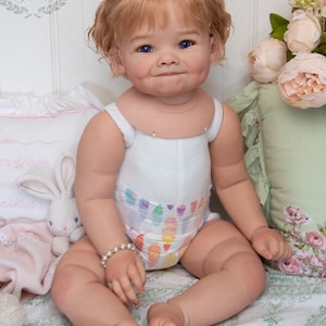 Pode incluir: Uma boneca de beb&ecirc; sorridente com cabelo loiro, olhos azuis e bochechas coradas est&aacute; vestindo um macac&atilde;o branco sem mangas com uma estampa colorida e um bracelete de p&eacute;rolas. A boneca est&aacute; sentada em um cobertor com padr&atilde;o floral.