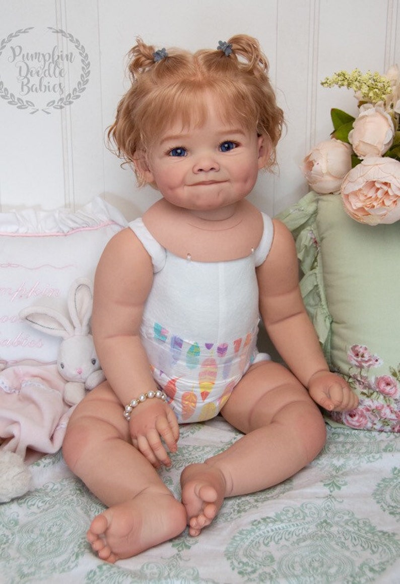 Pode incluir: Uma boneca de beb&ecirc; sorridente com cabelo loiro, olhos azuis e bochechas coradas est&aacute; vestindo um macac&atilde;o branco sem mangas com uma estampa colorida e um bracelete de p&eacute;rolas. A boneca est&aacute; sentada em um cobertor com padr&atilde;o floral.
