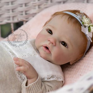 May include: A realistic baby doll with fair skin, red hair, and brown eyes, wearing a light-colored outfit with a white lace collar. The doll has a blue headband with white flowers and is lying on a pink blanket.