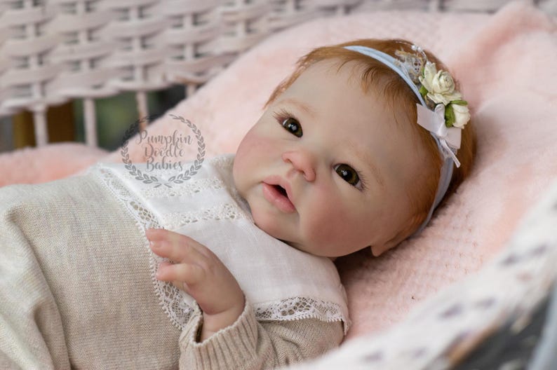 May include: A realistic baby doll with fair skin, red hair, and brown eyes, wearing a light-colored outfit with a white lace collar. The doll has a blue headband with white flowers and is lying on a pink blanket.