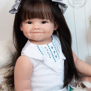 Pode incluir: Uma boneca de beb&ecirc; sorridente com cabelo preto comprido, vestindo uma camisa branca sem mangas com detalhes bordados, shorts verdes e la&ccedil;os prateados no cabelo. Ela est&aacute; segurando um coelho de pel&uacute;cia.