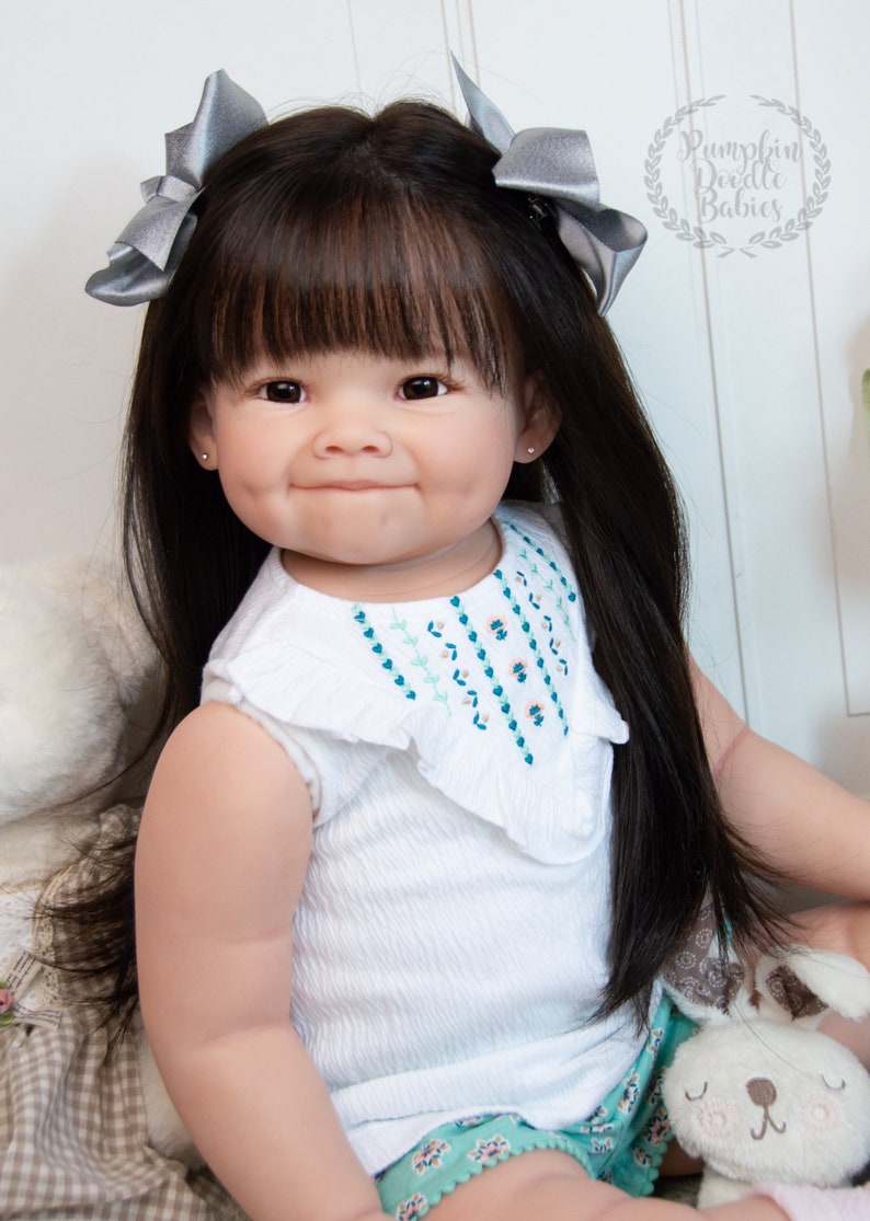 Pode incluir: Uma boneca de beb&ecirc; sorridente com cabelo preto comprido, vestindo uma camisa branca sem mangas com detalhes bordados, shorts verdes e la&ccedil;os prateados no cabelo. Ela est&aacute; segurando um coelho de pel&uacute;cia.