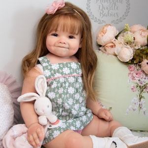 Pode incluir: Uma boneca realista de beb&ecirc; com cabelo castanho longo, vestindo um vestido floral verde e rosa, meias brancas e sapatos brancos. A boneca est&aacute; segurando um coelho de pel&uacute;cia branco e est&aacute; sentada em um travesseiro com padr&atilde;o floral branco e verde. A imagem inclui o texto "Pumpkin Doodle Babies".