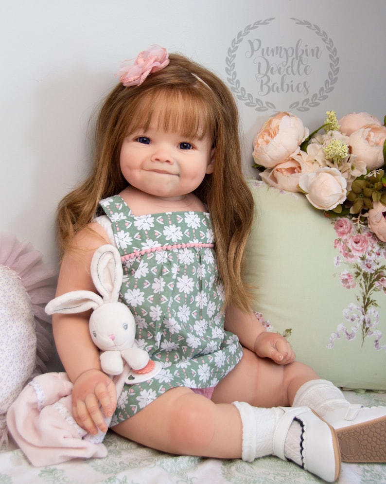 Pode incluir: Uma boneca realista de beb&ecirc; com cabelo castanho longo, vestindo um vestido floral verde e rosa, meias brancas e sapatos brancos. A boneca est&aacute; segurando um coelho de pel&uacute;cia branco e est&aacute; sentada em um travesseiro com padr&atilde;o floral branco e verde. A imagem inclui o texto "Pumpkin Doodle Babies".