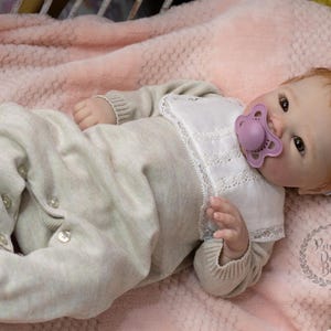 May include: A lifelike baby doll wearing a light beige knit onesie with button closures and a white bib. The doll has red hair, a pacifier, and a floral headband. The doll is lying on a pink blanket.