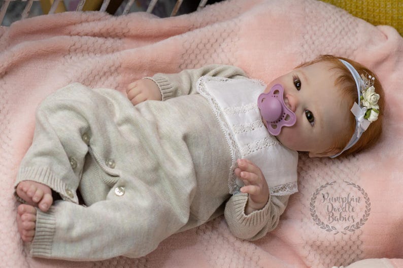 May include: A lifelike baby doll wearing a light beige knit onesie with button closures and a white bib. The doll has red hair, a pacifier, and a floral headband. The doll is lying on a pink blanket.