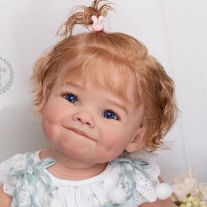Pode incluir: Um close-up de uma boneca beb&ecirc; com cabelo loiro encaracolado, olhos azuis e bochechas rosadas. A boneca est&aacute; vestindo um vestido branco com uma sobreposi&ccedil;&atilde;o de padr&atilde;o azul claro e branco. A boneca tem um pequeno prendedor de cabelo rosa com um design de coelho no cabelo.