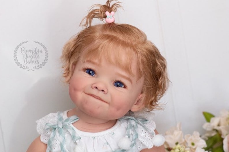 Pode incluir: Um close-up de uma boneca beb&ecirc; com cabelo loiro encaracolado, olhos azuis e bochechas rosadas. A boneca est&aacute; vestindo um vestido branco com uma sobreposi&ccedil;&atilde;o de padr&atilde;o azul claro e branco. A boneca tem um pequeno prendedor de cabelo rosa com um design de coelho no cabelo.