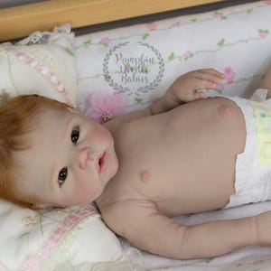 May include: A realistic baby doll with red hair and brown eyes, wearing a diaper, lying on a floral patterned blanket. The doll is positioned on a bed with a wooden frame. The text "Pumpkin Doodle Babies" is visible.