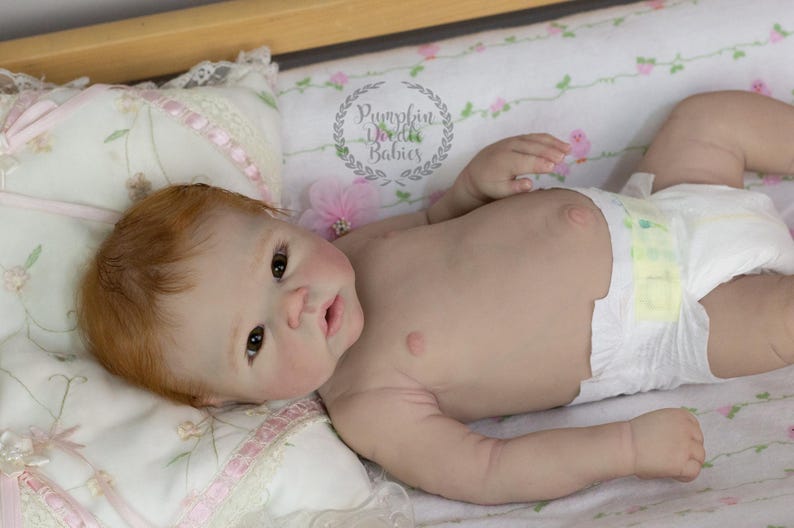 May include: A realistic baby doll with red hair and brown eyes, wearing a diaper, lying on a floral patterned blanket. The doll is positioned on a bed with a wooden frame. The text "Pumpkin Doodle Babies" is visible.